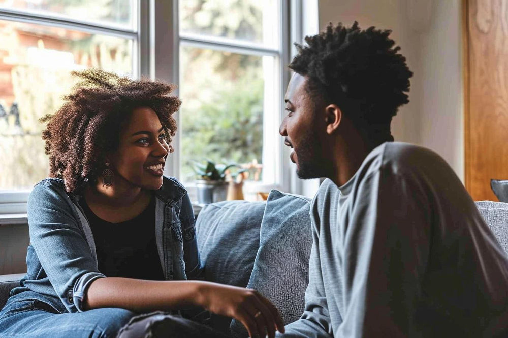 A couple is sitting on a couch talking in a room with large windows.
