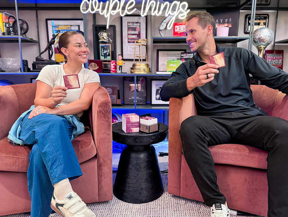 Shawn Johnson East & Andrew East in their podcast studio holding Couple Things cards and looking at each other