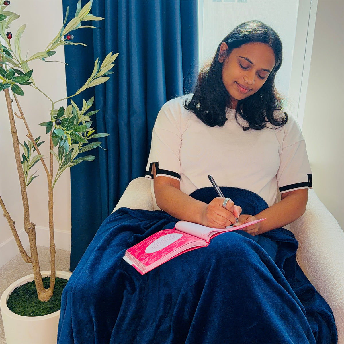Woman sitting on a chair with a blanket on her lap, writing in the Gratitude Prompted Journal 