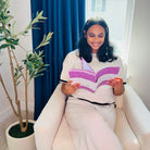 Woman sitting in a chair holding an Inspiration Prompted Journal with a plant and window in the background