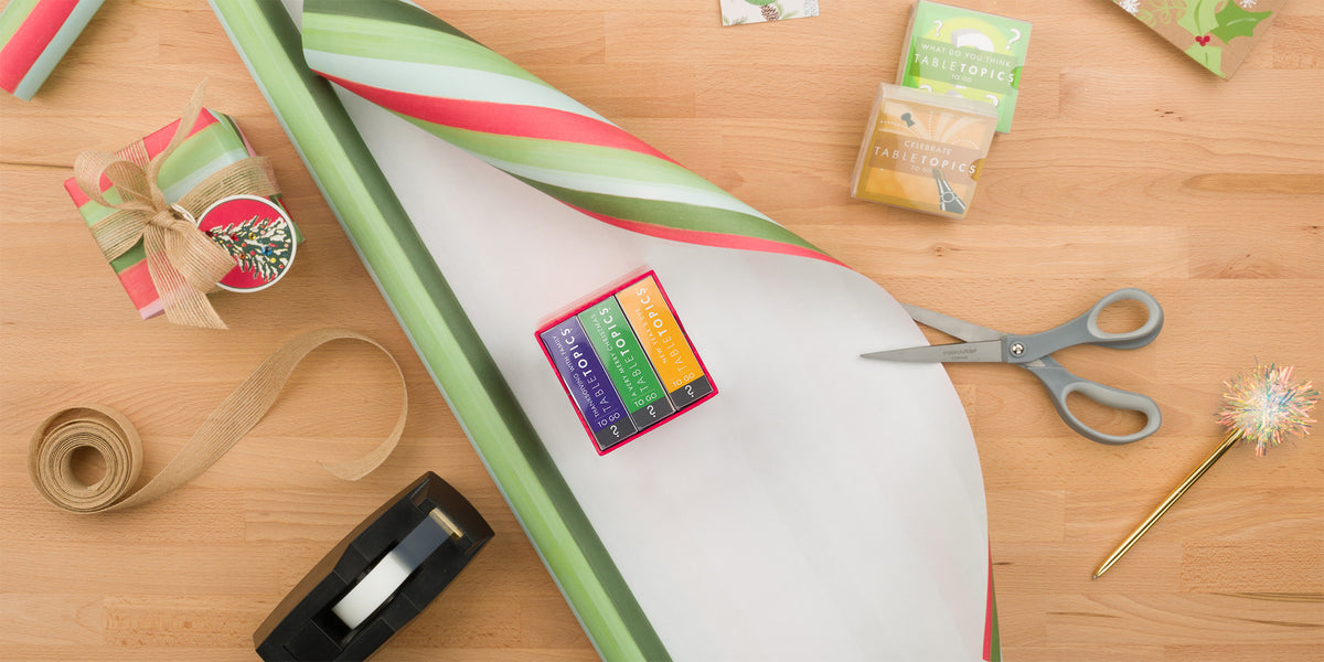 TableTopics Holiday Conversation Pack on top of wrapping paper with scissors, tape, and ribbon on table next to it