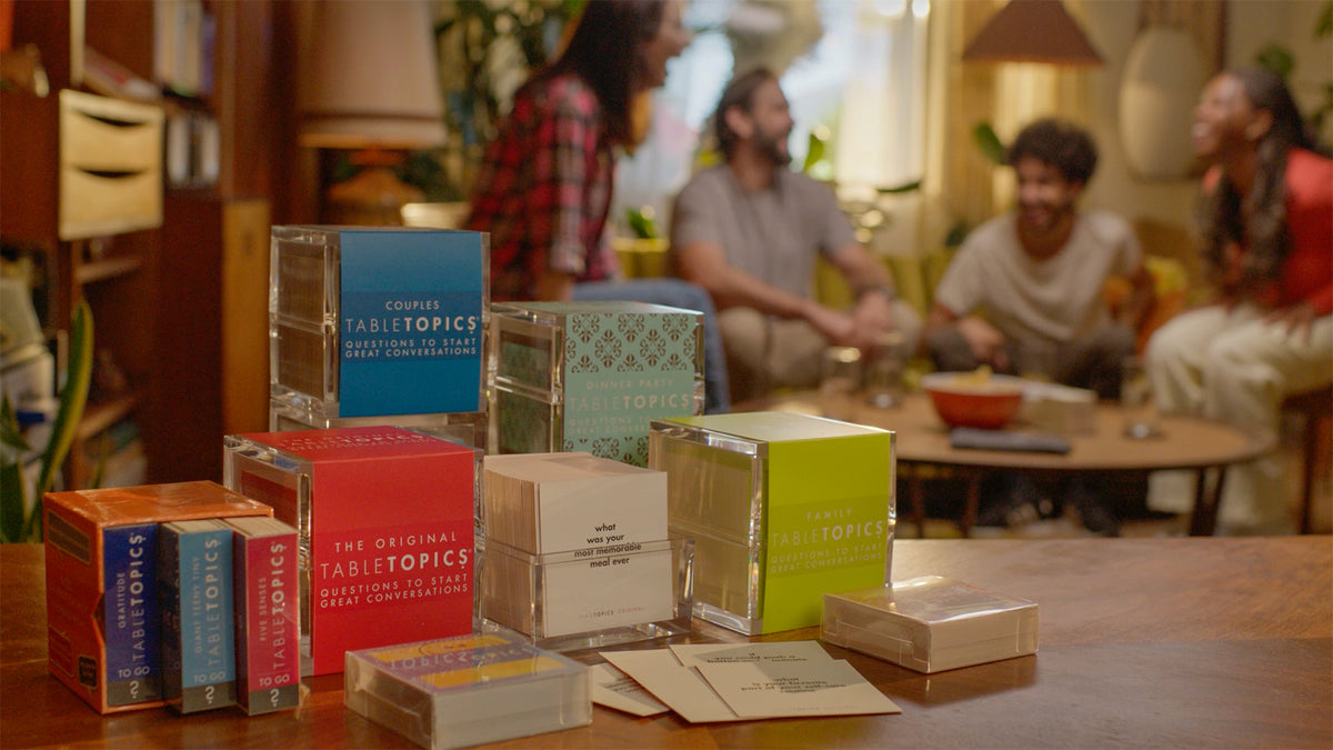 Friends playing TableTopics Original in living room in the background with a group of TableTopics sets in the foreground